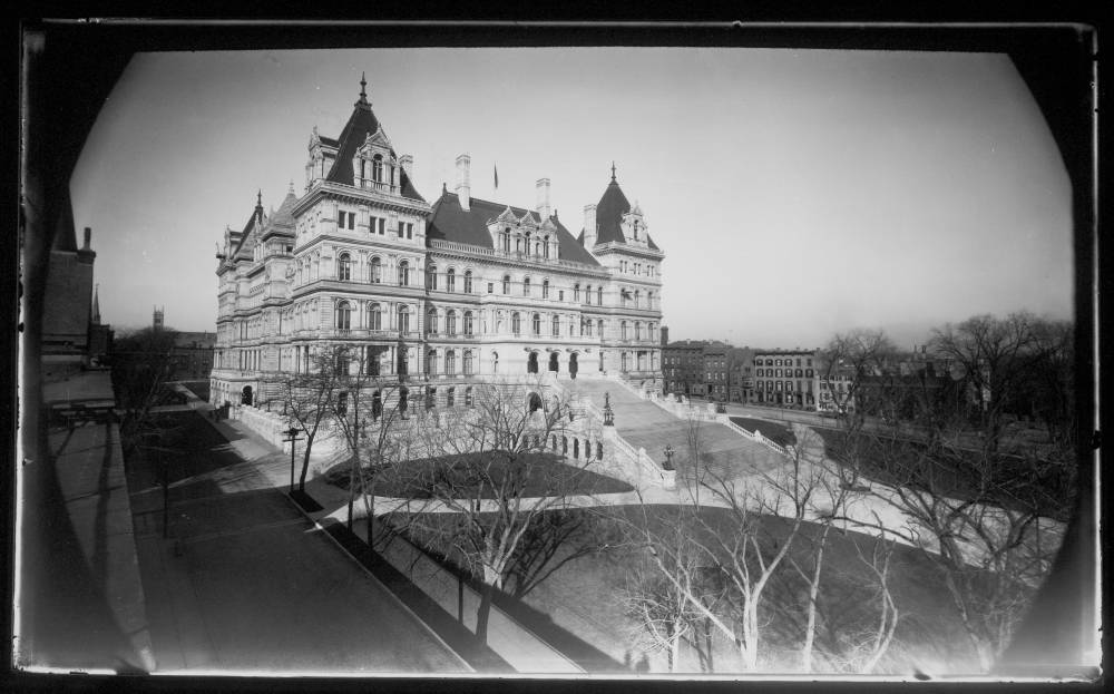 This black and white photograph of the New York State House features a grand and ornate building with a symmetrical facade, likely a government or historical structure, set against a clear sky. The building has a central tower and multiple smaller towers, each topped with pointed roofs. Wide, sweeping stairs lead up to the entrance, flanked by intricate stone railings. The surrounding area includes a paved walkway and manicured lawns with leafless trees, suggesting it is taken in late autumn or winter. In the background, smaller buildings and bare trees are visible, adding depth to the scene. The overall ambiance is majestic and formal.