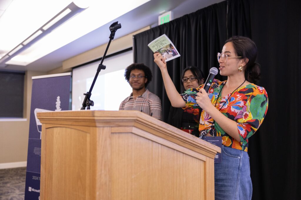 Jazzy holds up a copy of the Mapping for Equity zine and talks into the microphone about the project.