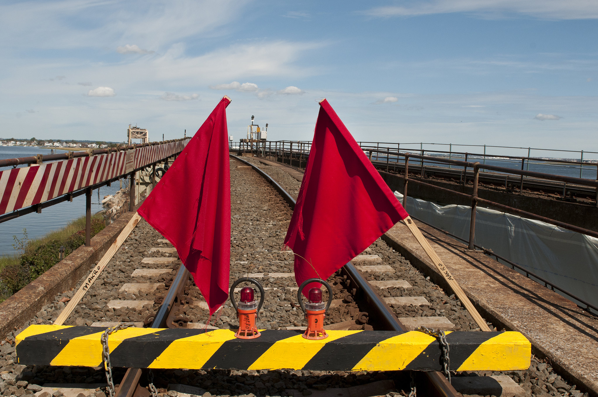 The image shows a railroad track blocked by a barrier with red flags and warning lights, set against a background of blue sky and distant buildings, indicating construction or maintenance.