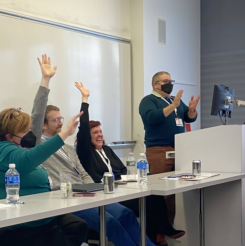 Three people sit at a table, raising their hands in response to a question posed by someone beside them, who stands behind a podium.