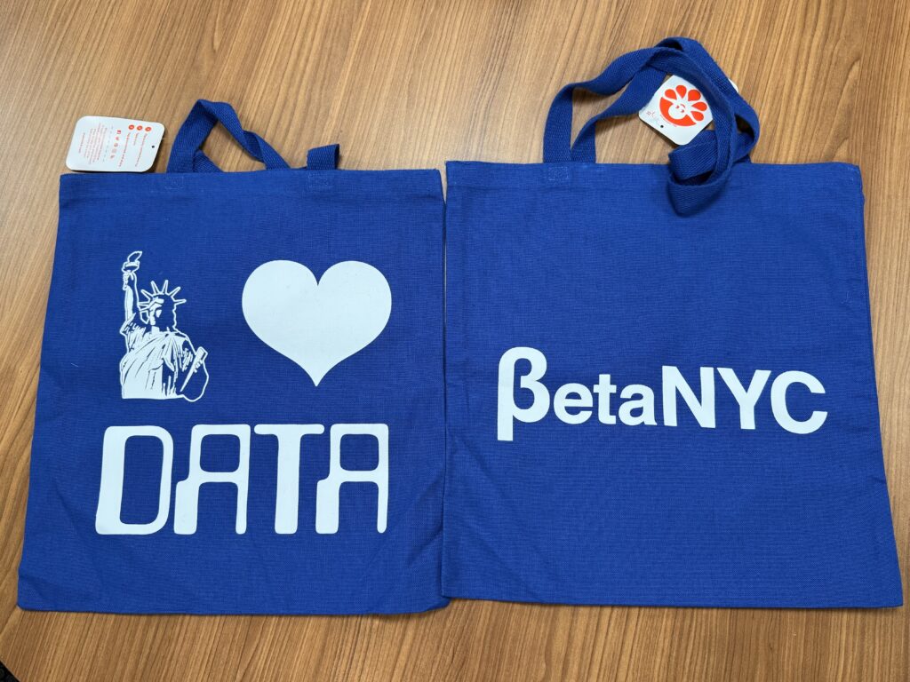 Two dark blue tote bags with a Statue of Liberty icon, a heart icon, and the words "Data" and "BetaNYC." The bags are on top of a wooden table.