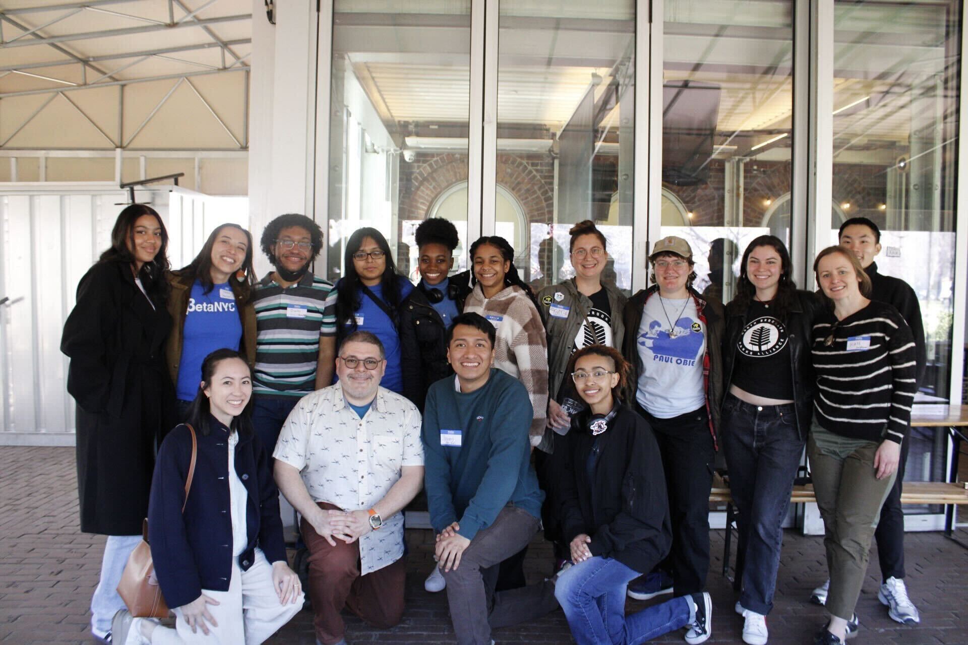 A group of fourteen people posing for a group photo in front of a glass-walled building. They are standing and kneeling, smiling and looking directly at the camera. Some are wearing BetaNYC branded shirts, while others have casual attire. The background shows an indoor space, and everyone appears to be gathered for an event or meeting. The atmosphere is friendly and welcoming.