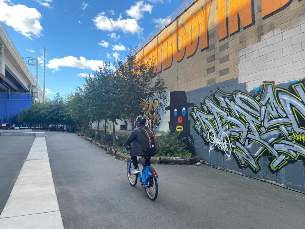 A person on a Citi Bike riding next to a colorful mural and trees.